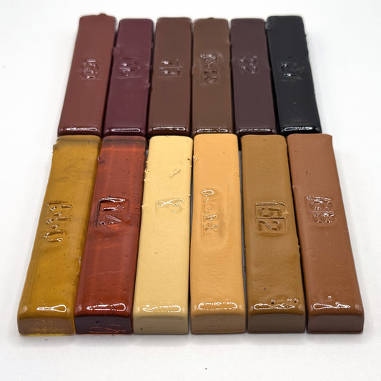 The Antique Dozen — twelve vintage hand-poured Merit burn-in half sticks arranged in two rows, antique furniture tones running from honey amber through burgundy and dark walnut to black