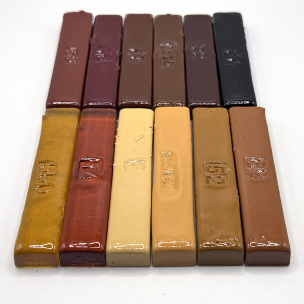 The Antique Dozen — twelve vintage hand-poured Merit burn-in half sticks arranged in two rows, antique furniture tones running from honey amber through burgundy and dark walnut to black